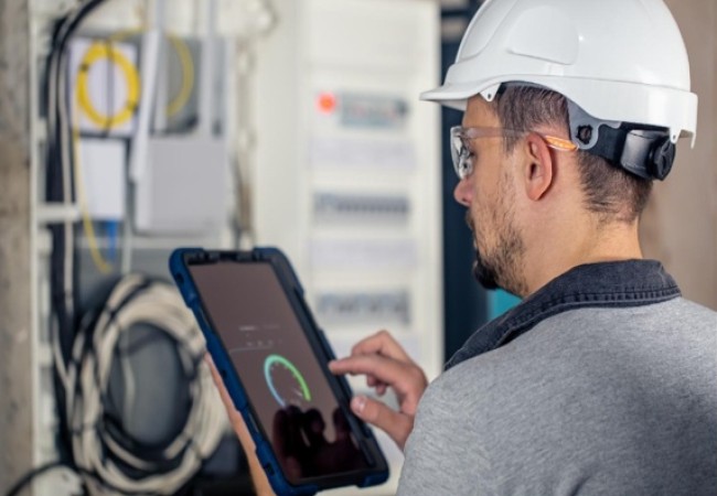 Electrical engineer using a tablet to inspect and monitor industrial electrical control panel equipment.